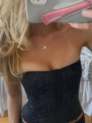 Svart blommig bustierkorsett - Säljer en svart axelbandslös korsett med blommigt mönster. Korsetten har hakknäppning framtill och en figurnära passform som framhäver midjan. Materialet är glansigt och ger en lyxig känsla. Perfekt att styla till jeans eller kjol.