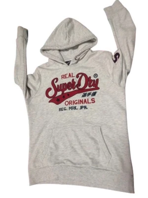 Ljusgrå Superdry hoodie med tryck - Snygg ljusgrå hoodie från Superdry med stor röd och blå logga på bröstet. Klassisk känguruficka framtill och huva med dragsko. Tillverkad i mjukt bomullsmaterial som är skönt att ha på sig. Perfekt för en avslappnad streetwear-look.