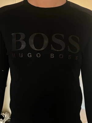 Svart långärmad tröja Hugo Boss - Snygg svart långärmad tröja från Hugo Boss med stor logga i svart på bröstet. Tröjan har rund halsringning och är tillverkad i ett mjukt material som sitter skönt på kroppen. Perfekt för en stilren och modern look.
