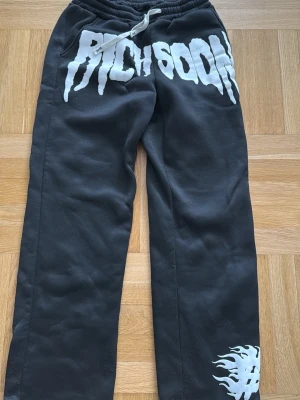 Svarta mjukisbyxor Rich Soon - Svarta mjukisbyxor från Rich Soon med vit dragsko i midjan och stora vita tryck fram, inklusive loggan och en eldflamma vid benet. Byxorna har avslappnad passform och är tillverkade i mjukt material, perfekta för chill dagar.