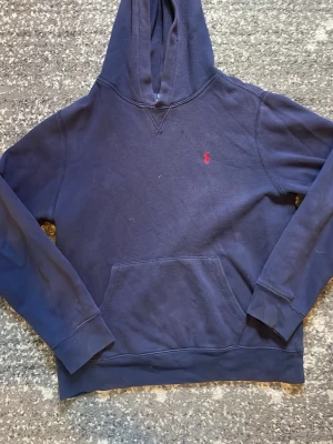 Mörkblå hoodie från Polo Ralph Lauren - Mörkblå hoodie från Polo Ralph Lauren med klassisk röd logga broderad på bröstet. Hoodien har huva, känguruficka och ribbade muddar vid ärmslut och nederkant. Tillverkad i mjukt bomullsmaterial som är skönt att bära. Storlek 14-16 år är som xs. Mycket bra skick