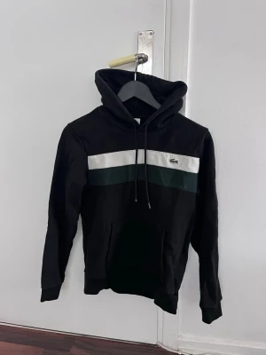 Svart hoodie från Lacoste med ränder - Snygg svart hoodie från Lacoste med vita och mörkgröna ränder över bröstet och klassiska krokodilloggan. Hoodien har huva med dragsko och en stor magficka. Tillverkad i mjukt bomullsmaterial, perfekt för en chill och sportig look.