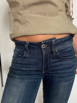 Mörkblå bootcut jeans - Säljer ett par mörkblå bootcut jeans med klassisk femficksdesign och kontrastsömmar. Jeansen har låg midja och lätt slitningar framtill för en snygg vintagekänsla. Perfekta till sneakers eller boots.