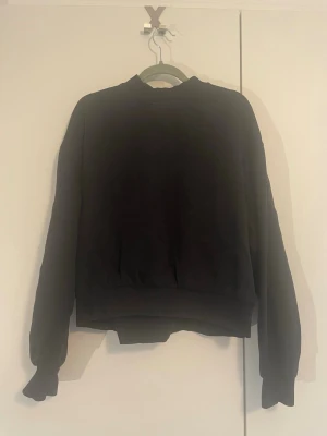 Svart sweatshirt - En enkel svart sweatshirt med rund hals och ribbade muddar vid ärmslut och nederkant. Tröjan har en avslappnad passform och är tillverkad i mjukt bomullsmaterial. 