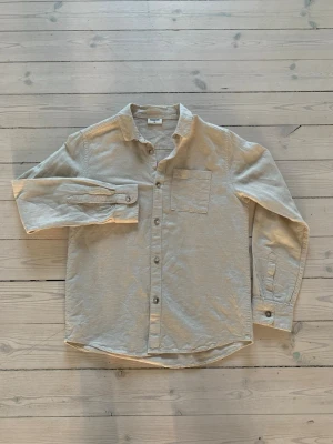 Beige linneskjorta från Asket - Säljer en stilren beige skjorta från Asket i linne. Skjortan har klassisk krage, bröstficka och knappar framtill. Perfekt för en avslappnad men snygg look. Långärmad modell med enkel, tidlös design.