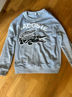 Lacoste crew neck  - Snygg grå sweatshirt från Lacoste med stort tryck av krokodil och logga på bröstet. Tröjan har rund halsringning, långa ärmar och ribbade muddar. Perfekt för en avslappnad och trendig stil.