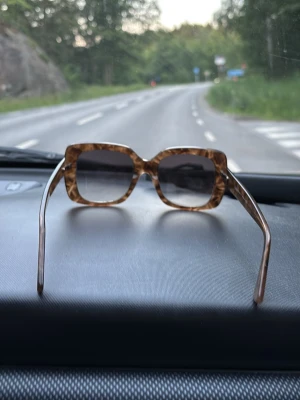 Bruna fyrkantiga solglasögon från Glas - Säljer ett par trendiga solglasögon från Glas med fyrkantiga bågar i brunmelerad plast. Snygg retrostil som passar perfekt till sommaren. Glasen är mörka och bågarna har en cool mönstrad finish.