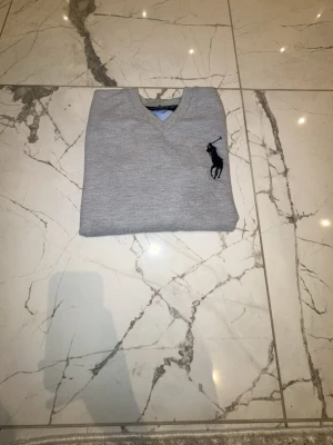 Grå tröja med V-ringning från Polo Ralph Lauren - Snygg grå tröja med V-ringning från Polo Ralph Lauren. Tröjan har ett svart broderat polospelar-märke på bröstet och ribbade muddar vid ärmslut och nederkant. Perfekt för en stilren och avslappnad look.