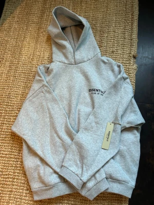  Fear of God Essentials hoodie Grå M - Snygg grå hoodie från Fear of God Essentials med svart logga på bröstet och ryggen. Klassisk oversized passform, mjukt material och stor huva. Perfekt för en avslappnad streetwear-look. Dragsko saknas för en clean vibe.