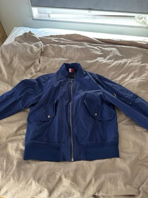 Marinblå bomberjacka Tommy Hilfiger - Snygg marinblå bomberjacka från Tommy Hilfiger med dragkedja framtill, två stora fickor med lock och tryckknappar samt ribbade muddar. Insidan har foder i rött, vitt och blått. Klassisk college-look och perfekt till streetwear.