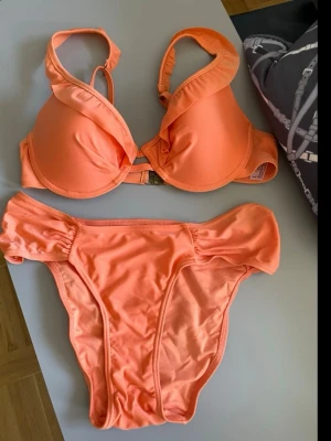Orange bikini med volangdetaljer - Snygg orange bikini med vadderad bygel-bh och volangdetaljer på axelbanden. Matchande bikiniunderdel med rynkade sidor för extra stil. Perfekt för strandhäng eller poolparty!