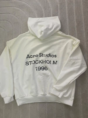 Vit hoodie Acne Studios Stockholm - Snygg vit hoodie från Acne Studios med tryck på ryggen där det står 'Acne Studios STOCKHOLM 1996'. Hoodien har en klassisk passform med huva och ribbade muddar. Perfekt för en clean och stilren look. Materialet känns mjukt och skönt mot huden.