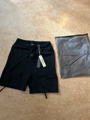 Svarta shorts från Essentials Fear of God - Svarta shorts från Essentials Fear of God med broderad logga på benet. De har elastisk midja med snörning och är tillverkade i mjuk bomull. Nya i förpackning