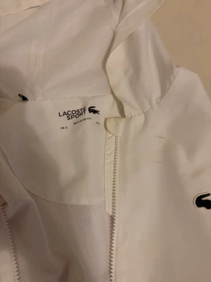 Vit vindjacka från Lacoste Sport - Vit vindjacka från Lacoste Sport med huva och dragkedja. Jackan har svarta ränder längs nederkanten och det klassiska Lacoste-krokodil-logotypen på bröstet. Materialet är lätt och andas, perfekt för att hålla vinden ute. Passar ej längre. Mycket bra skick