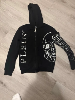 Svart hoodie från Philipp Plein Junior - Snygg svart hoodie med dragkedja från Philipp Plein Junior. Stora silverfärgade tryck med texten 'PLEIN' på ena sidan och en cool döskalle på bröstet samt grafiskt mönster på ärmen. Tillverkad i mjukt bomullsmaterial och har huva.