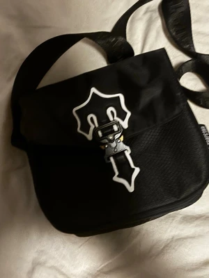 Svart axelväska från Trapstar - Säljer en svart axelväska från Trapstar med justerbar rem och coolt vitt emblem på locket. Väskan har en unik spänndetalj framtill och är gjord i slitstarkt tygmaterial. Perfekt för dig som vill ha en streetstyle-vibe och plats för det viktigaste.