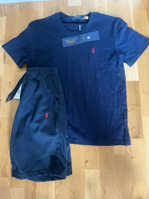 Mörkblå set från Polo Ralph Lauren - Snyggt mörkblått set från Polo Ralph Lauren med t-shirt och shorts. Båda plaggen har den klassiska röda logotypen broderad på bröstet och benet. T-shirten är i mjuk bomull och shortsen har resår i midjan med dragsko för skön passform.
