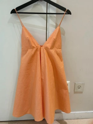  Trendig orange kort klänning från Zara  - Säljer en söt kort klänning med tunna axelband och v-ringning. Klänningen är orange med vita smala ränder och har en luftig, a-linjeformad passform. Perfekt för varma dagar och enkel att styla med sneakers eller sandaler.