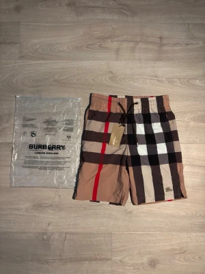 Burberry badbyxor med rutigt mönster - Säljer ett par snygga Burberry badbyxor med klassiskt beige, svart, vitt och rött rutmönster. De har elastisk midja med dragsko och en bakficka med knapp. Materialet är lätt och snabbtorkande, perfekt för stranden eller poolen. 