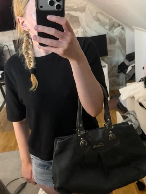 Svart handväska från Coach - Snygg svart handväska från Coach med dubbla axelband i läder och silverfärgade metalldetaljer. Väskan har diskret mönster med Coach-loggor i tyget och en metallogo framtill. Perfekt storlek för att få plats med allt du behöver.