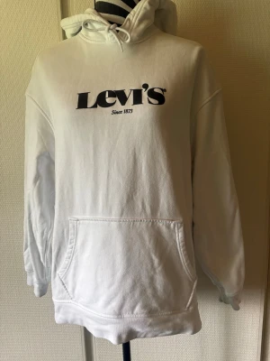 Vit hoodie från Levi's med logga - Säljer en vit hoodie från Levi's med stor svart logga och texten 'Since 1873' på bröstet. Hoodien har huva med dragsko, stor magficka och ribbade muddar vid ärmar och nederkant. Perfekt för en avslappnad och stilren look.