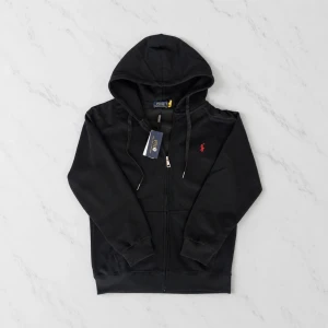Svart hoodie från Polo Ralph Lauren - Snygg svart hoodie från Polo Ralph Lauren med dragkedja framtill och klassisk huva. Den har en liten röd logga broderad på bröstet och ribbade muddar vid ärmar och nederkant. Perfekt för en chill och stilren look. Storlek M men rekommenderar för dom som har S.