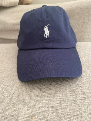 Mörkblå keps från Polo Ralph Lauren - Snygg mörkblå keps från Polo Ralph Lauren med klassisk vit broderad logga framtill och 'POLO' broderat bak. Justerbar rem baktill för perfekt passform. Tillverkad i bomullstwill som ger en stilren och sportig look.
