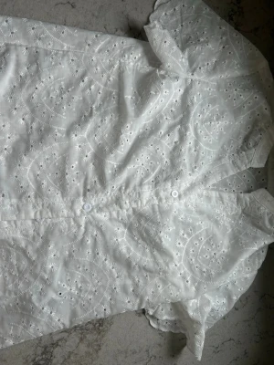 Vit broderad blus med kort ärm - Söt vit blus med broderade blommor och knappar framtill. Blusen har korta ärmar med vågig kant och är gjord i ett tunt, luftigt bomullstyg. Perfekt för en somrig och romantisk look.