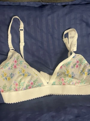 Blommig bralette från H&M - Söt bralette från H&M i vitt meshmaterial med blommigt mönster i pastellfärger. Modellen har justerbara axelband och bred resår under bysten för extra komfort. Perfekt för dig som gillar en mjuk och lätt bh med romantisk känsla.