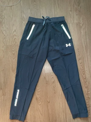 gråa träningsbyxor från Under Armour - Snygga svarta träningsbyxor från Under Armour med vita detaljer och logga på benet. Byxorna har resår i midjan med dragsko, två fickor med dragkedja och smal passform. Perfekta för gymmet eller löpning.