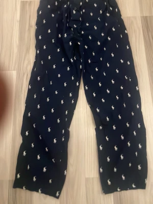 Mörkblå pyjamasbyxor Polo Ralph Lauren - Säljer ett par mörkblå pyjamasbyxor från Polo Ralph Lauren med vitt broderat logomönster över hela byxan. Byxorna har resår i midjan och dragsko för bästa passform. Perfekta för chillkvällar hemma. Knappt använda, kan även tänka mig gå ner i pris!