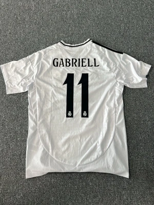 Real Madrid matchtröja Adidas vit - Säljer en vit Real Madrid matchtröja från Adidas med svarta detaljer och klubbmärke på bröstet. Tröjan har trycket 'Gabriell' och nummer 11 på ryggen samt 'Emirates Fly Better' på framsidan. Perfekt för fotbollsfans som vill sticka ut på planen.
