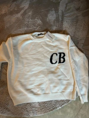 Vit stickad tröja CB Cole Buxton  - Vit stickad tröja från Cole Buxton med svarta bokstäver 'CB' på bröstet. Tröjan har rund halsringning, ribbade muddar och är tillverkad i mjukt material. Enkel och stilren design som passar till mycket.