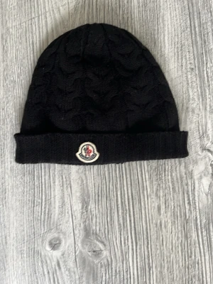 Svart kabelstickad mössa från Moncler - Snygg svart kabelstickad mössa från Moncler med uppvikt kant och klassisk Moncler-logga framtill. Mössan är tillverkad i mjukt garn och har ett stilrent mönster som ger extra struktur och känsla. QR koden och chipen går att scanna. Denna mössan är äkta från moncler. 