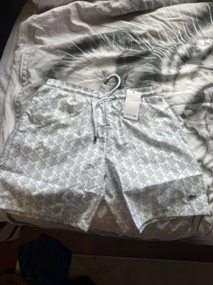 Vita badbyxor - Säljer ett par vita badbyxor med grått geometriskt mönster och dragsko i midjan. Klassisk passform och diskret Lacoste-logga på benet. Perfekta för stranden eller poolen. Passar även M