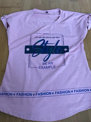 Ljusrosa t-shirt med tryck - Trendig ljusrosa t-shirt med blått texttryck 'Style' och dekorativt band med texten 'FASHION' längst ner. T-shirten har korta ärmar med knappdetalj på axlarna och rund halsringning. Perfekt för dig som gillar en avslappnad och cool look.