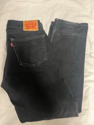 Svarta Levi's jeans - Säljer ett par klassiska svarta Levi's 501 jeans i rak modell. Jeansen har fem fickor, knappgylf och den ikoniska röda Levi's-lappen på bakfickan. Tillverkade i slitstark denim av bomull. Perfekta för dig som gillar tidlös och clean stil. Köpta för 1000kr