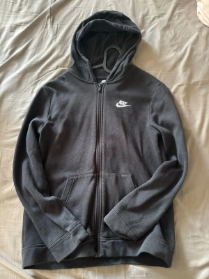 Svart Nike zip hoodie  - Säljer en svart hoodie från Nike. Hoodien har dragkedja framtill, två fickor och en klassisk huva. Nike-loggan är broderad i vitt på bröstet. Materialet är mjukt och skönt, perfekt för chill dagar eller när du vill ha en sportig look.