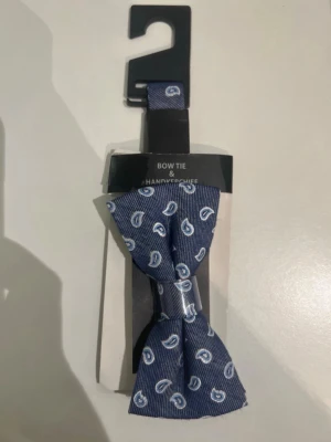 Blå fluga med paisleymönster Cubus - Snygg blå fluga från Cubus med vitt paisleymönster. Tillverkad i 61% polyester och 39% viskos, perfekt för att ge din outfit en stilren touch. Levereras i förpackning tillsammans med matchande näsduk.