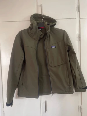 Olivgrön vindjacka från Patagonia - Snygg olivgrön vindjacka från Patagonia med huva och dragkedja. Jackan har en bröstficka med dragkedja, två sidofickor och är fodrad med mjuk fleece på insidan. Perfekt för blåsiga dagar och outdoor-äventyr.