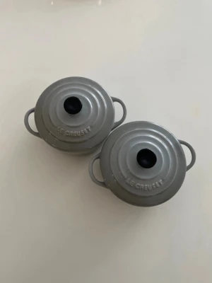 Grå minigrytor från Le Creuset - Två små grå minigrytor från Le Creuset i keramik med lock och svarta knoppar. Perfekta för servering eller små portioner. Grytorna har rund form och handtag på båda sidor. Insidan är ljusbeige och utsidan är blankt grå.