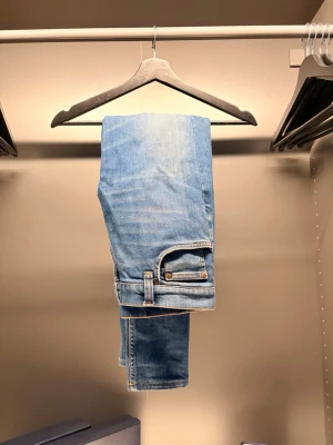  Nudie Jeans - Säljer ett par ljusblå jeans från Nudie Jeans med klassisk femficksdesign och kontrastsömmar. Jeansen har en avslappnad passform och är tillverkade i mjuk denim av bomull. Bakfickan har Nudie Jeans karakteristiska broderi.