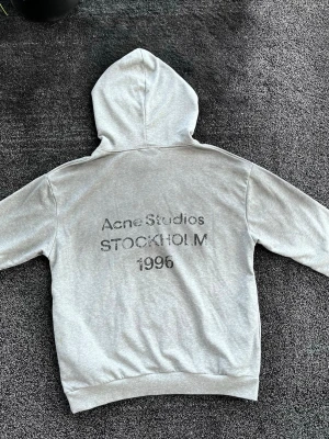 Acne Studios hoodie - Säljer en stilren grå hoodie från Acne Studios med tryck på ryggen där det står 'Acne Studios STOCKHOLM 1996'. Hoodien har huva, ribbade muddar och är gjord i mjukt bomullsmaterial. Perfekt för dig som gillar minimalistisk design och streetwear. Storlek L, skick 9/10. KÖP GÄRNA BUNDLE (20%rabatt på allt!!!)