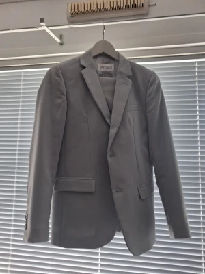 Ljusgrå kavaj från Opposuits - Snygg ljusgrå kavaj från Opposuits med klassisk passform och två knappar framtill. Kavajen har smala slag, tre fickor och knappar vid ärmsluten. Perfekt för dig som vill ha en stilren och modern look. Hör av dig vid frågor