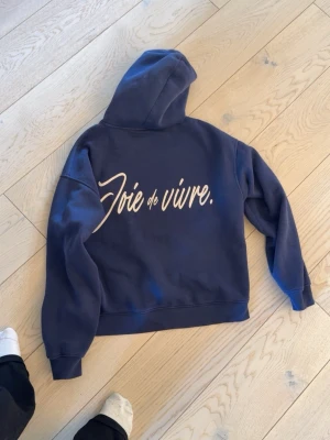 Mörkblå hoodie med texttryck - Säljer en mörkblå hoodie med stor vit text 'Joie de vivre.' på ryggen. Hoodien har en klassisk huva och en stor magficka framtill. Tillverkad i mjukt bomullsmaterial och har ribbade muddar vid ärmslut och nederkant. Perfekt för en chill och avslappnad stil.