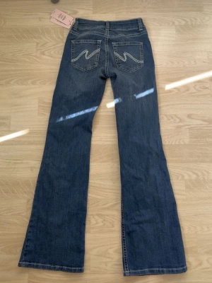 Blå bootcut jeans från Nelly  - Säljer ett par blå bootcut jeans från nelly med klassisk femficksmodell och snygga broderade detaljer på bakfickorna. Jeansen har normal midja och är tillverkade i ett mjukt denimtyg med lätt stretch för skön passform. Dom är helt nya men tyvärr är de ett litet hål ovanför bakfickorna, något som inte syns så mycket eller som är enkelt att laga.