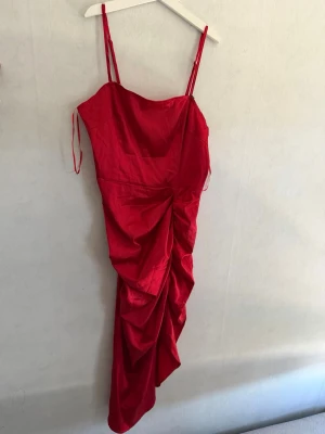 Röd långklänning med drapering - Snygg röd långklänning med smala axelband och draperad detalj framtill. Klänningen har en figurnära passform och är tillverkad i ett glansigt material som ger en lyxig känsla. Perfekt för dig som vill sticka ut.