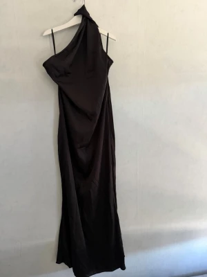 Svart oneshoulder långklänning - Elegant svart långklänning med oneshoulder-design och smala axelband. Klänningen har en mjuk, följsam passform och ett stilrent uttryck som passar perfekt till speciella tillfällen. Materialet har en lätt glansig yta för extra lyxig känsla.