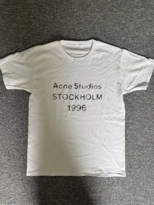 Vit Acne Studios t-shirt Stockholm - Vit t-shirt från Acne Studios med tryck 'STOCKHOLM 1996' på bröstet. Klassisk passform och rund halsringning. Perfekt basic-plagg med stilren design och diskret logga framtill.  ALLA lappar saknas därav priset 