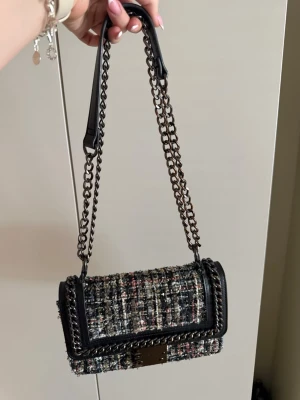 Axelväska i tweed med kedjerem - Snygg axelväska från Carvela i svart, grått och rosa tweed med detaljer i svart skinn och en kraftig kedjerem. Väskan har en rektangulär form och stängs med ett metallspänne framtill. Perfekt för dig som vill ha en trendig och unik accessoar.